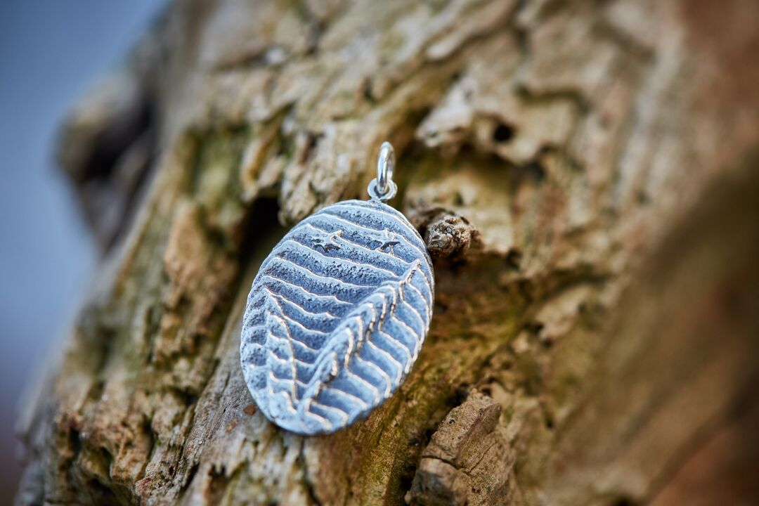 Detail of cuttlebone cast pendant of carved river rushes in silver set against the bark of a tree.