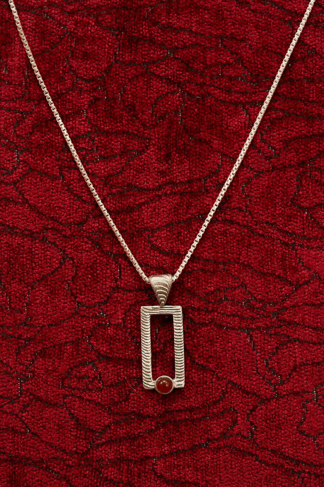 Cuttlebone cast rectangular framed pendant and necklace in gold set against rich dark red fabric.